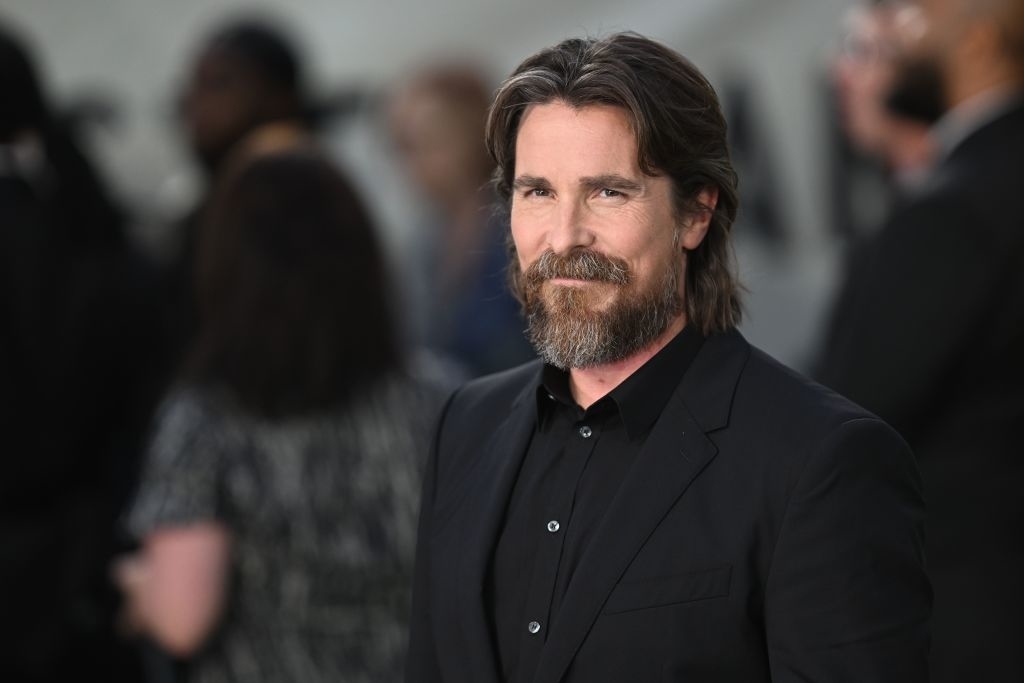 Christian Bale in a black suit with long hair and beard smiling at an event