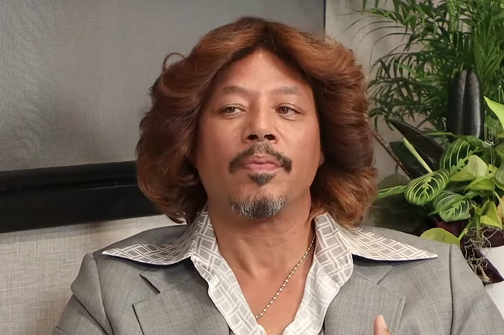 Man with medium-length wavy hair sitting, wearing a striped suit jacket and a patterned shirt