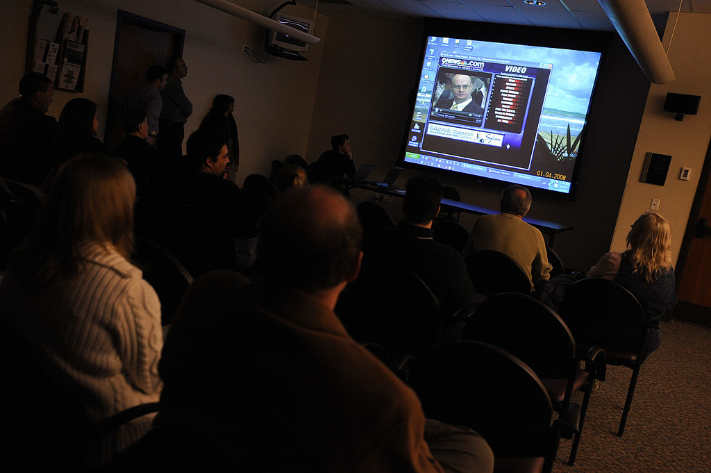 A group of people watching a presentation in a dark room with a screen displaying a slide