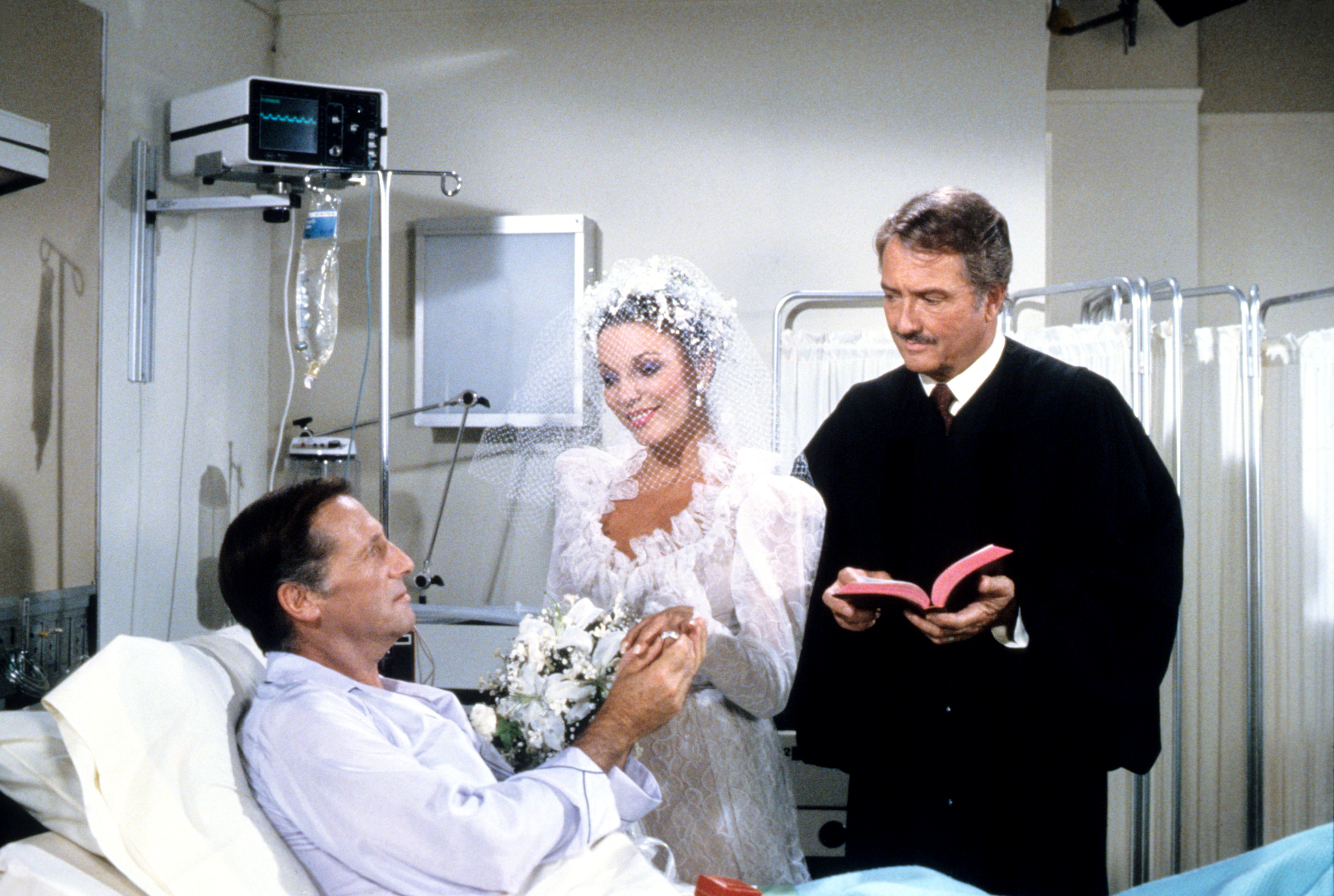 A bride in a full gown and a man in a suit stand by a patient in a hospital bed, officiating a wedding