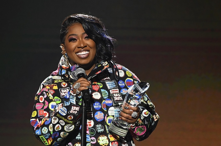 Missy Elliott wearing a patch-covered jacket, holding a microphone and award
