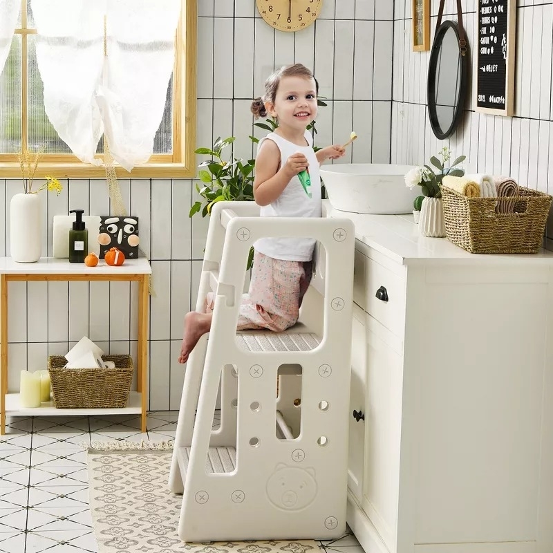 Child stands on a white step stool at bathroom sink, holding a toothbrush. Stool has side safety rails, suitable for kids
