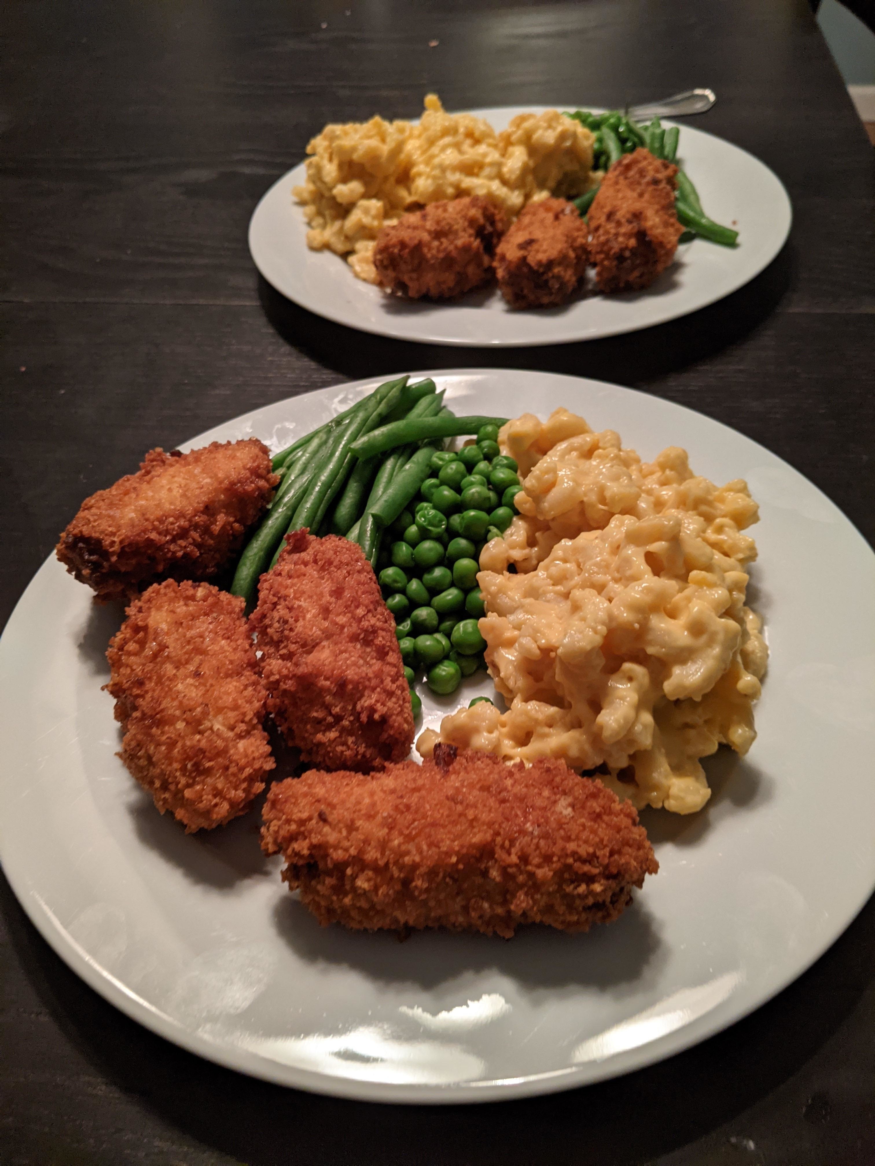 Two plates of food with breaded chicken, green beans, peas, and macaroni and cheese