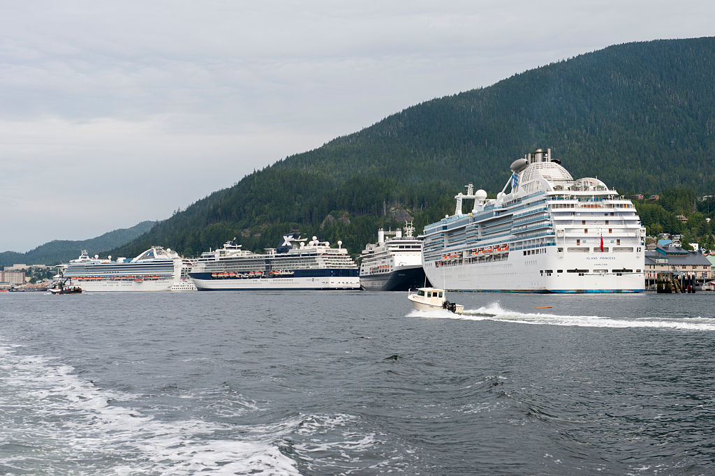 Three large cruise ships docked in a harbor with a forest-covered hill in the background