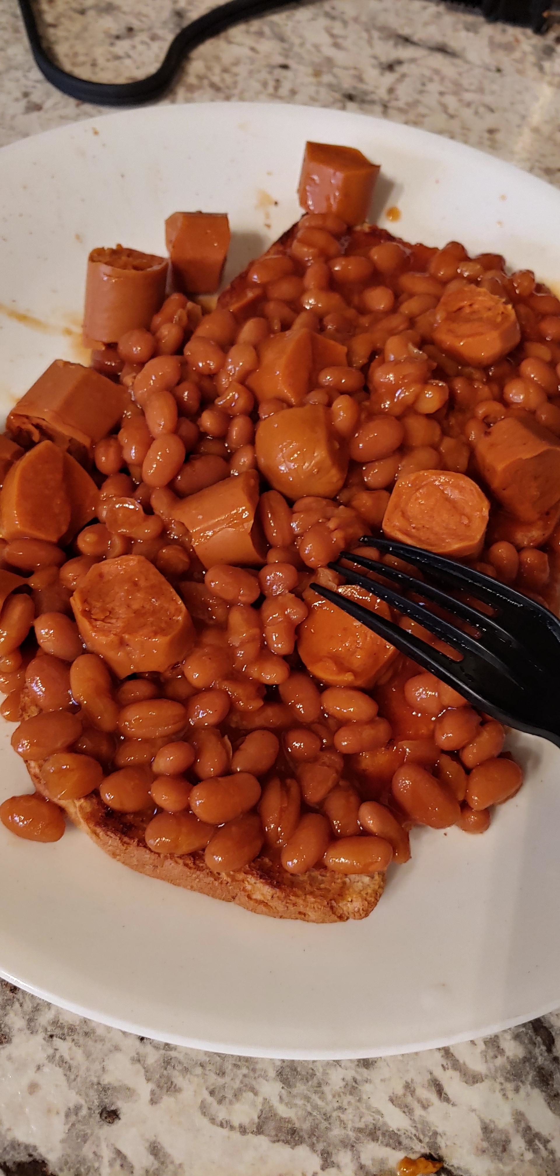 A plate of baked beans and sliced hot dogs with a black fork