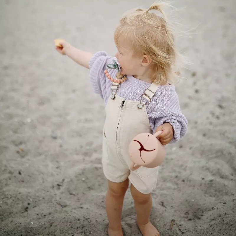 Toddler holding a toy at the beach, wearing overalls with a plush bear bag