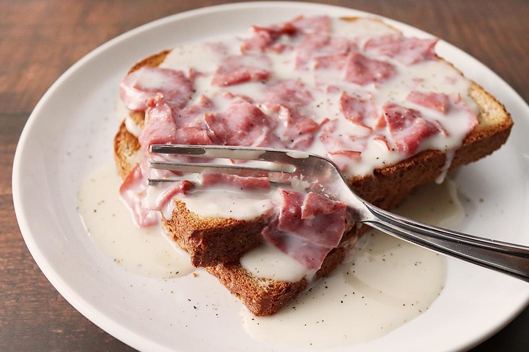 Slice of bread topped with cream sauce and ham, on a plate with a fork resting on top