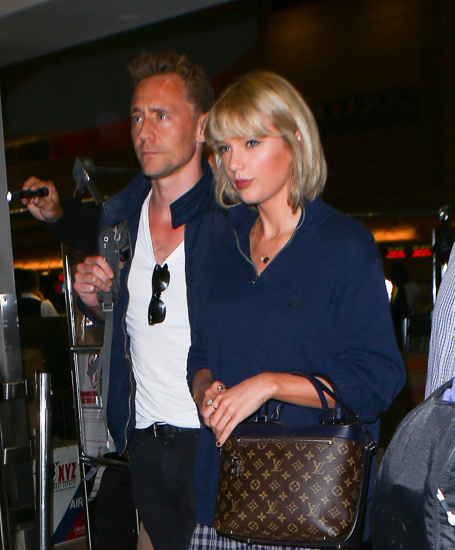 Taylor Swift in a navy top and plaid skirt with a shoulder bag, walking with Tom Hiddleston in a white shirt