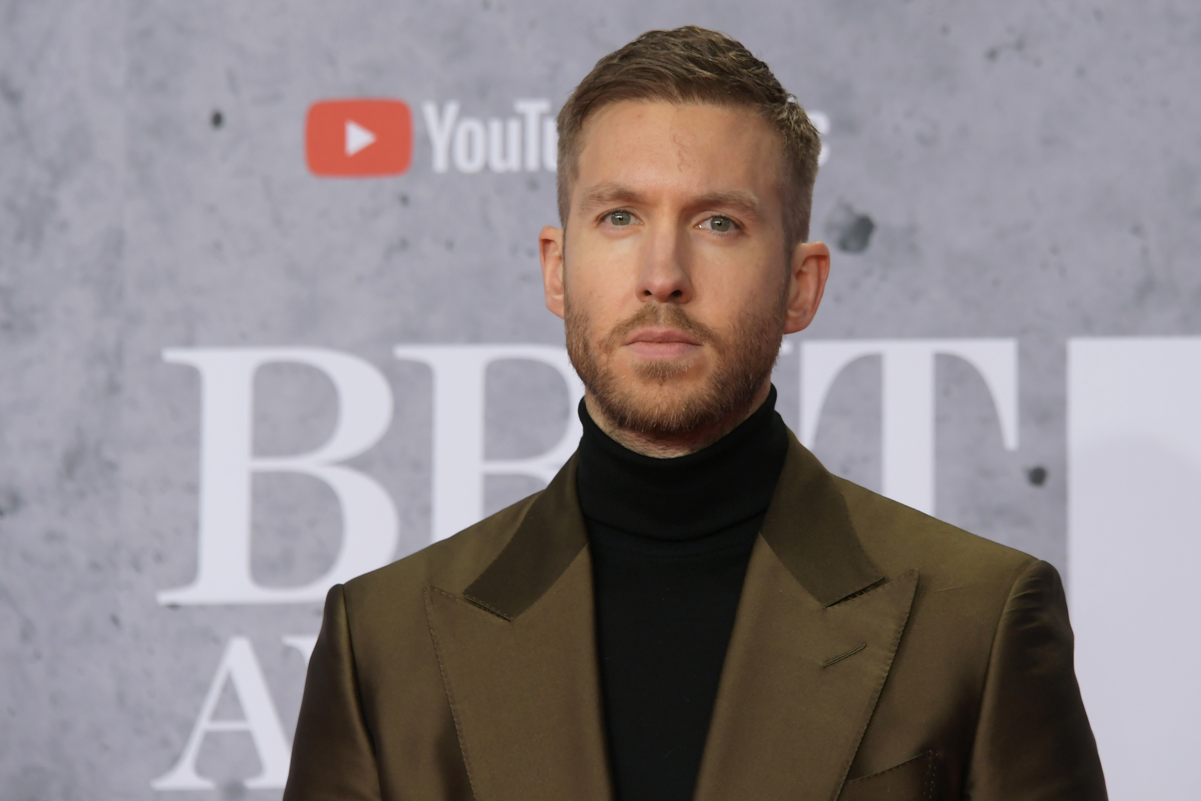 Calvin Harris in a black turtleneck and brown suit jacket standing in front of a backdrop with logos