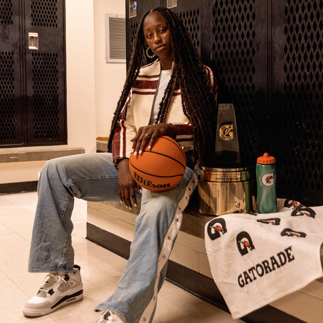 Person seated with a basketball, styled in casual attire, beside Gatorade bottles and a towel, in a locker room setting