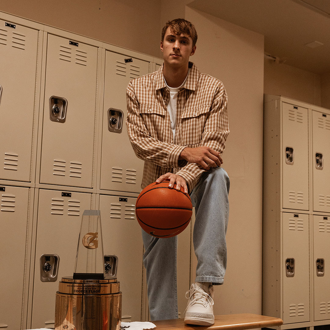 Person in a checkered jacket and jeans sitting on a bench holding a basketball, with lockers in the background