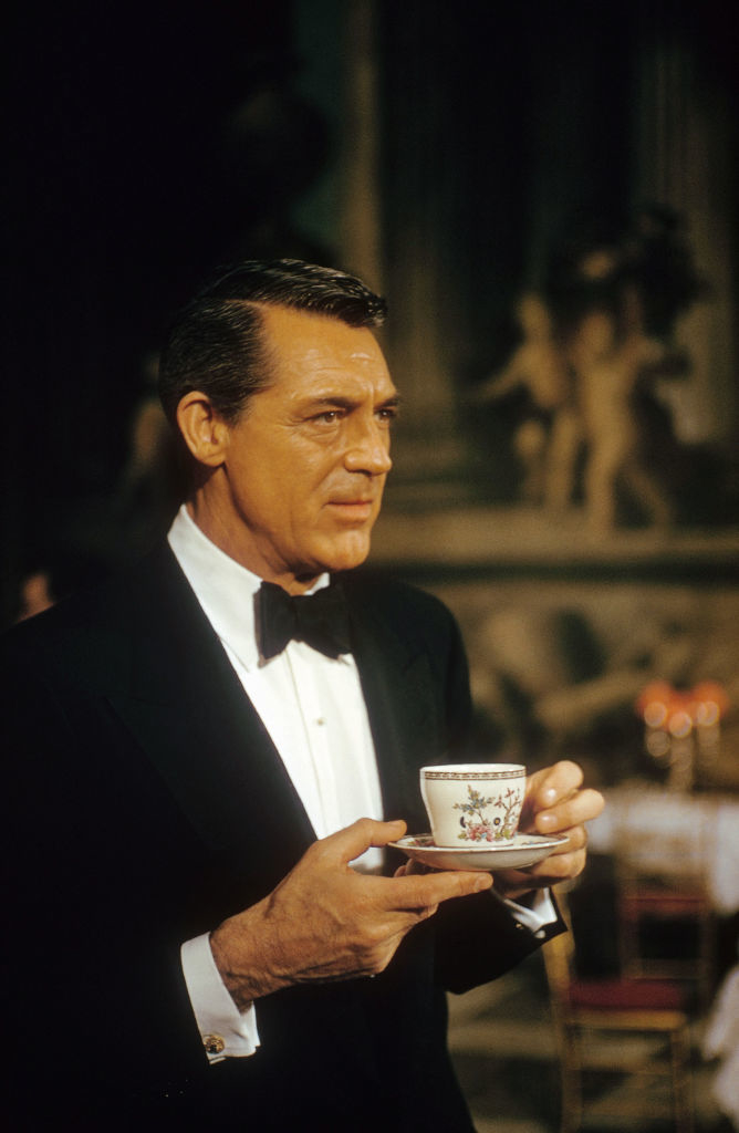 Cary in tuxedo holds a teacup, poised and elegant, against an ornate backdrop