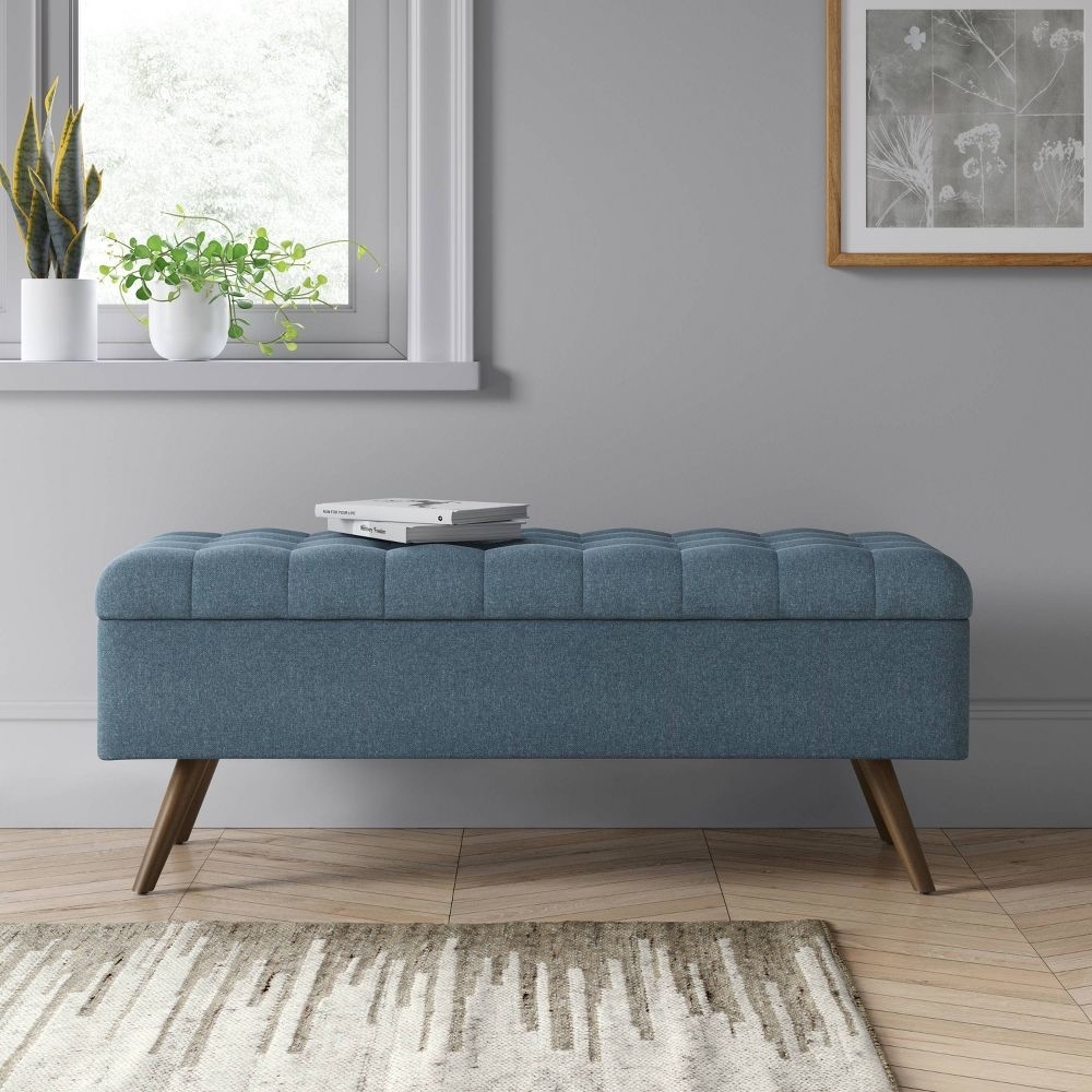 A tufted blue ottoman with storage, angled wooden legs, placed in a minimalist living room setting
