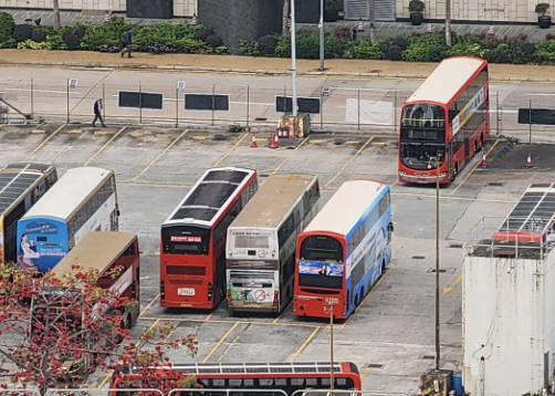 Multiple buses parked in a lot, some double-decker, with a city backdrop