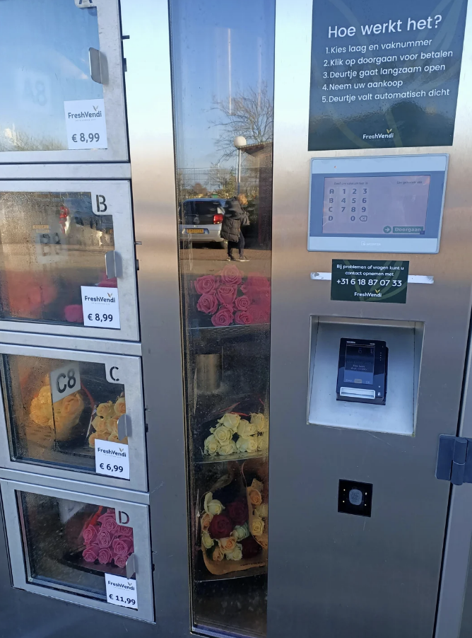 Vending machine with various flowers for sale, instructions on use to the right, reflection of two people visible