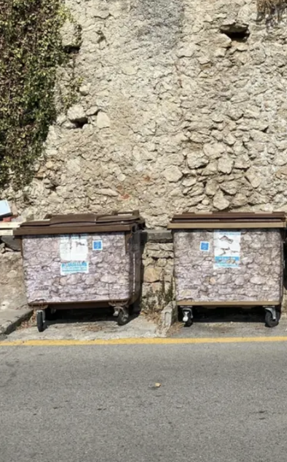 Two dumpsters against a stone wall, with visible posters on them