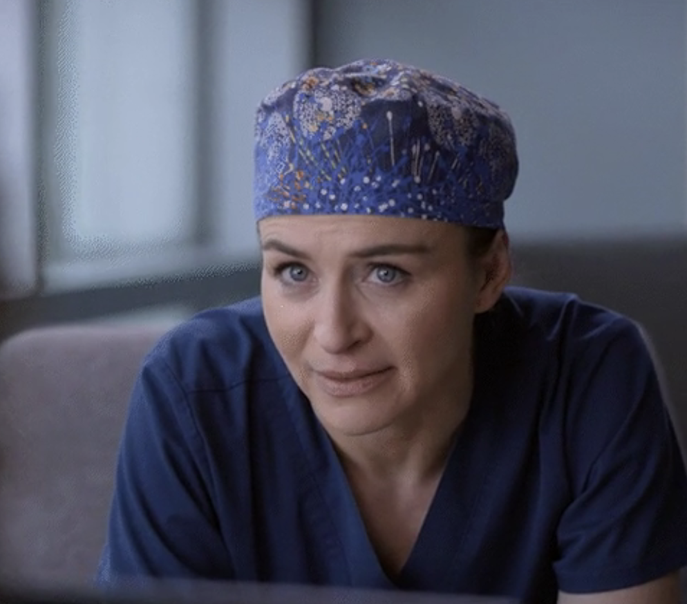A medical professional in scrubs and a patterned scrub cap looks thoughtfully towards the camera