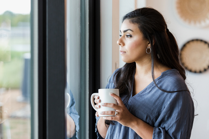 Woman in a casual top holding a mug and looking out a window