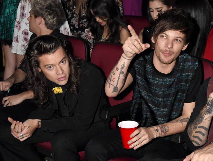 Harry Styles in a suit with a flower pin and Louis Tomlinson in a striped tee seated at an event, talking