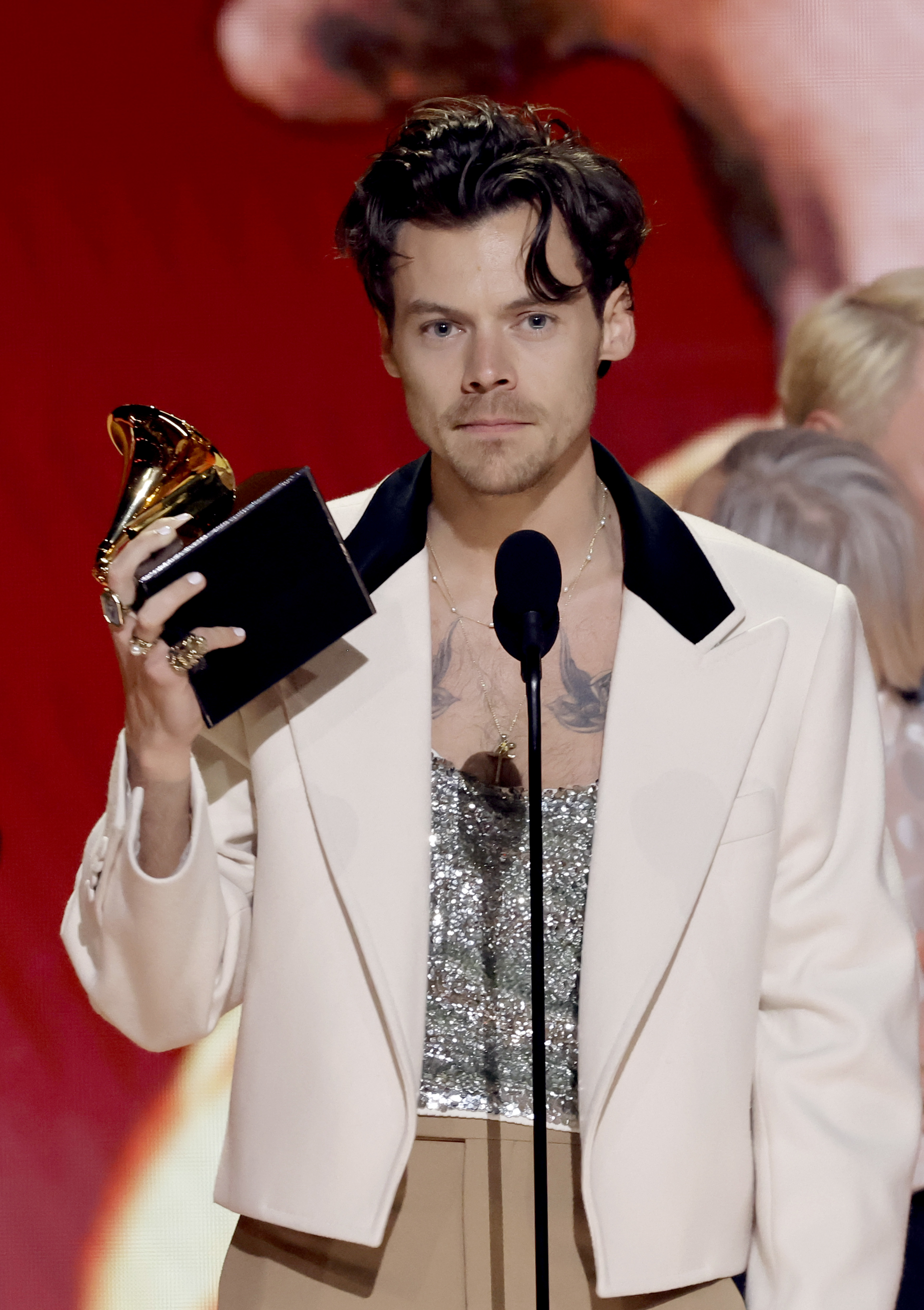Harry Styles in a jacket and sparkly top holding a Grammy award on stage