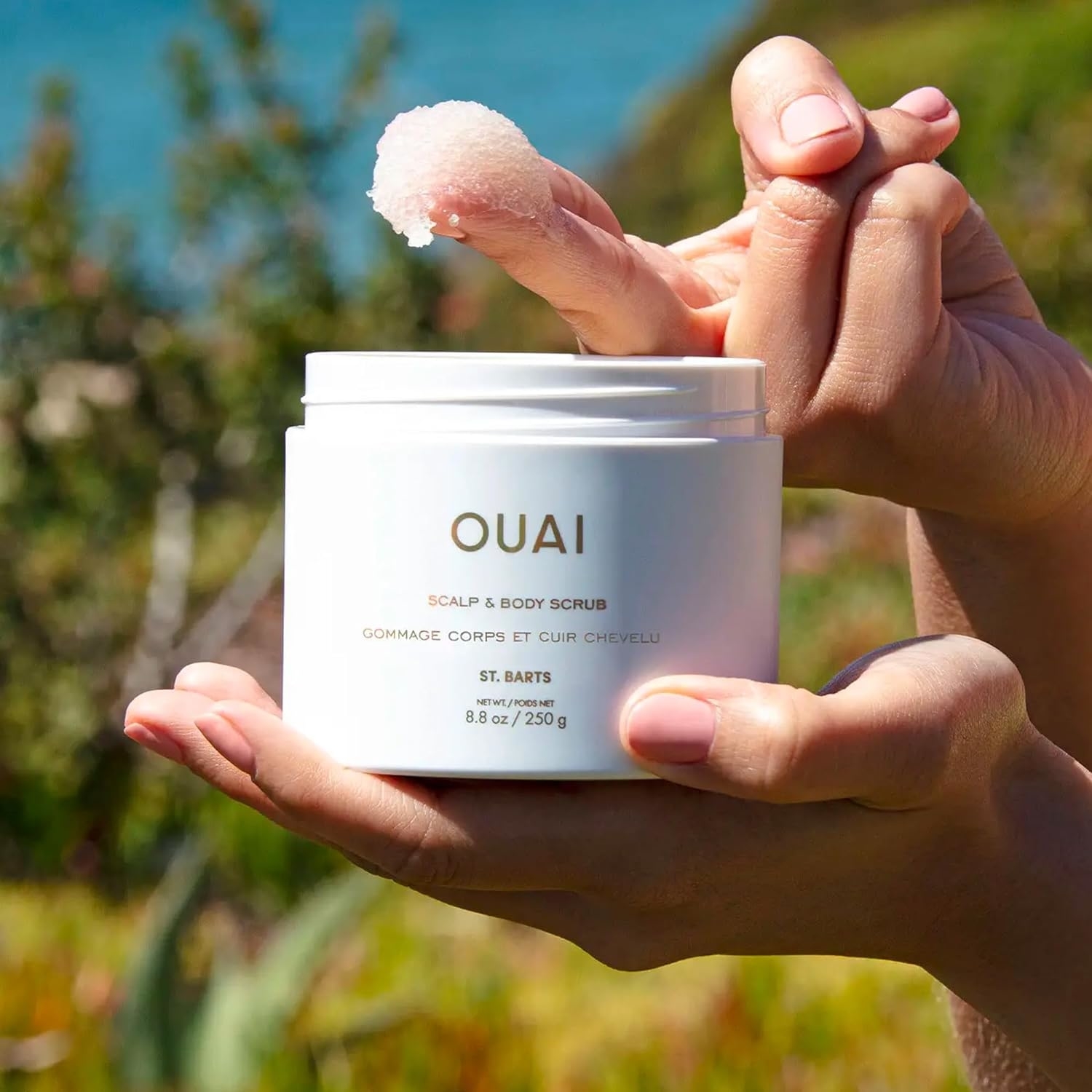 Person holding a jar of OUAI scalp and body scrub with a dab of product on their finger against a natural backdrop