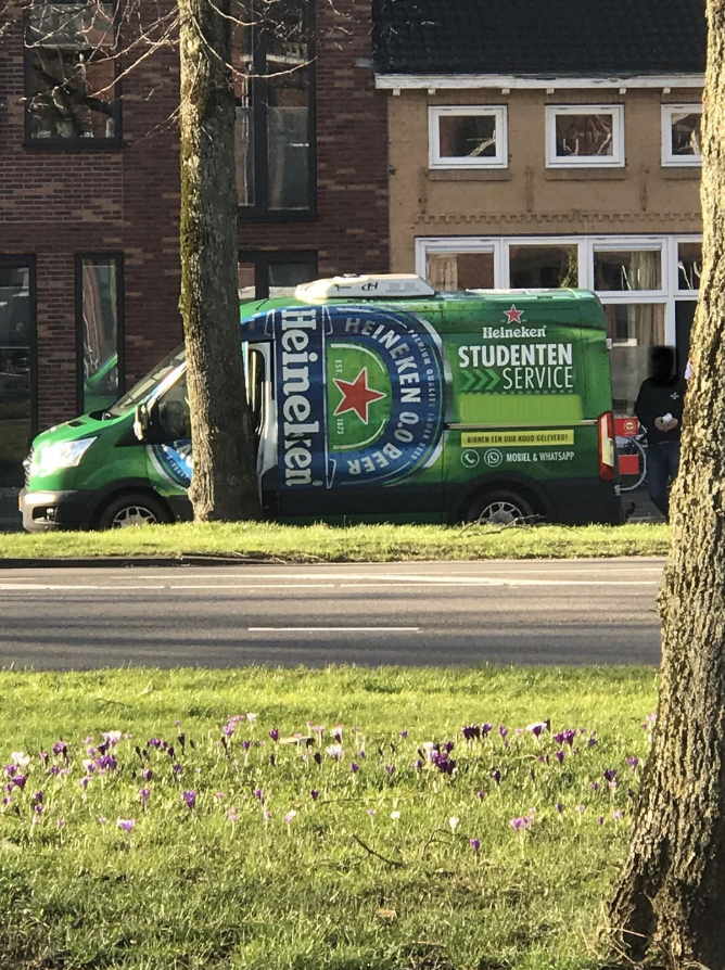 A Heineken-branded van with &quot;Student Service&quot; text, parked by a tree-lined street, near some flowering plants