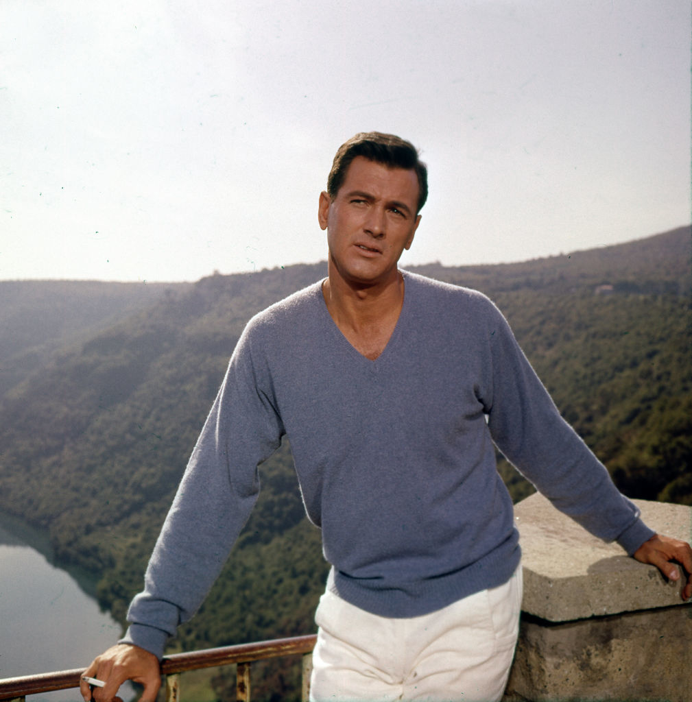 Rock Hudson poses for a portrait wearing a sweater in front of a mountainscape