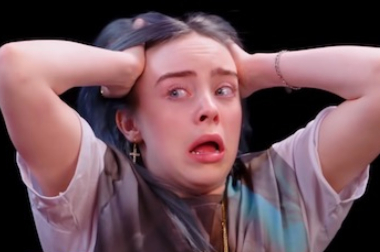 Billie Eilish with a shocked expression, hands on head, wearing a beige top