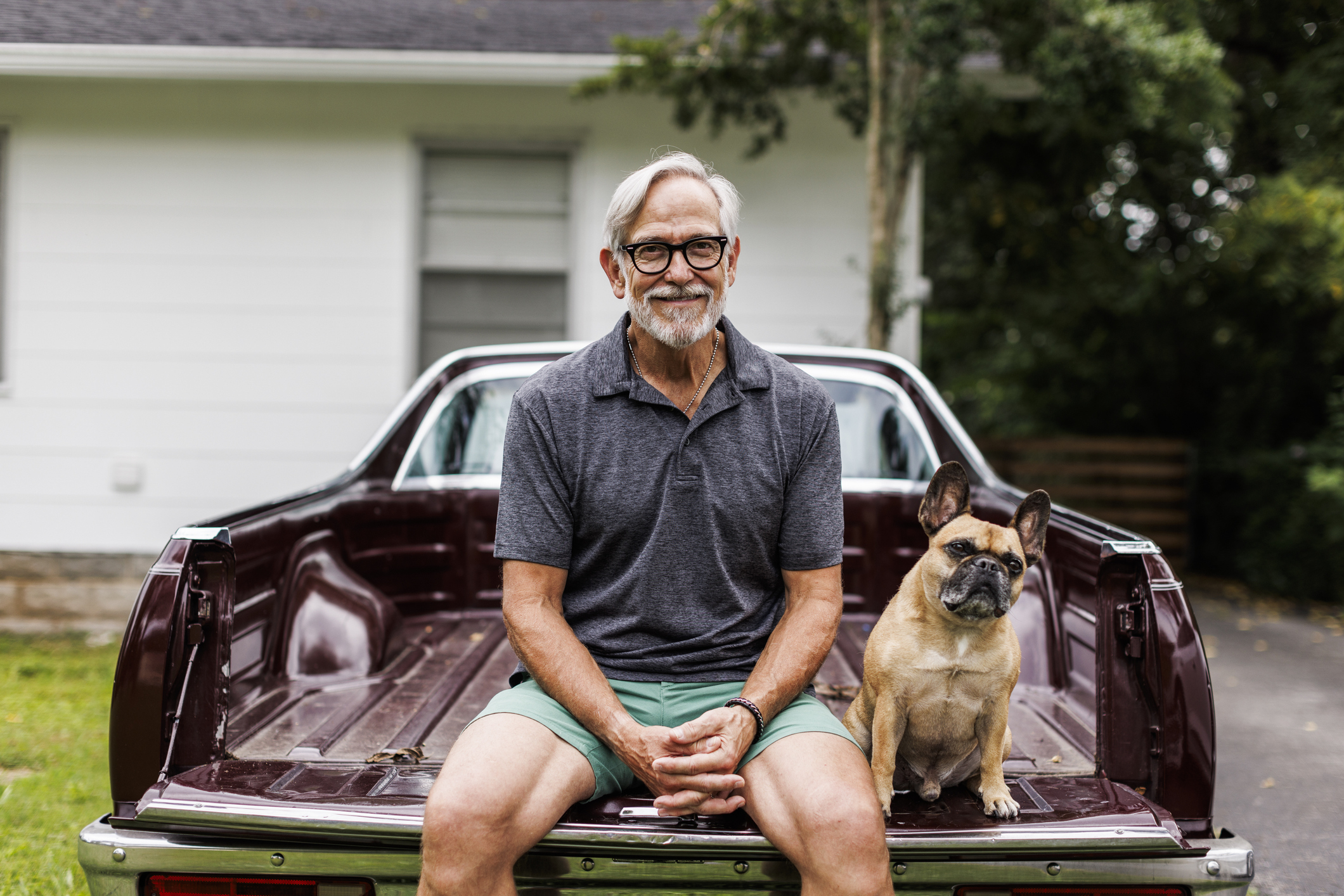 Smiling man and French Bulldog sitting on the trunk of a vintage car with a house in the background