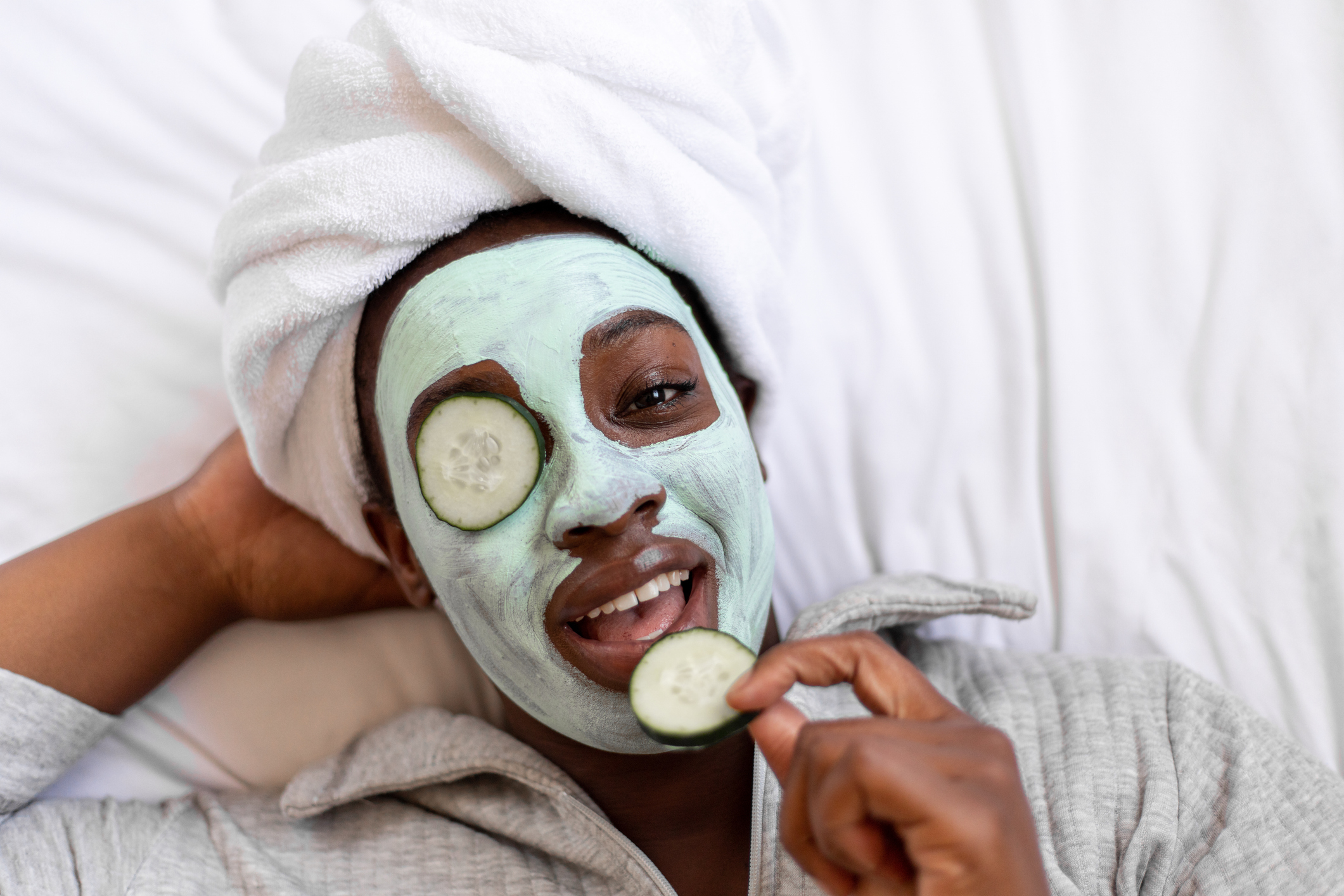 Person with a face mask and towel on head, lying down with cucumber slices on eyes, promoting skincare relaxation