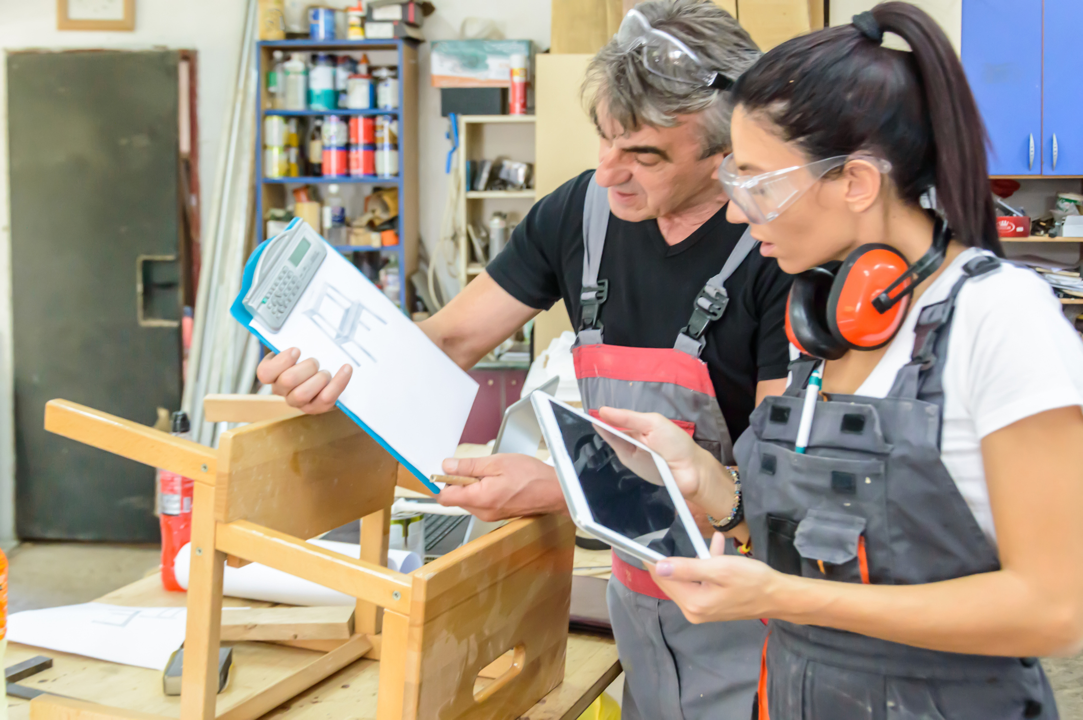 Two carpenters, one holding plans, discuss work at a woodworking bench