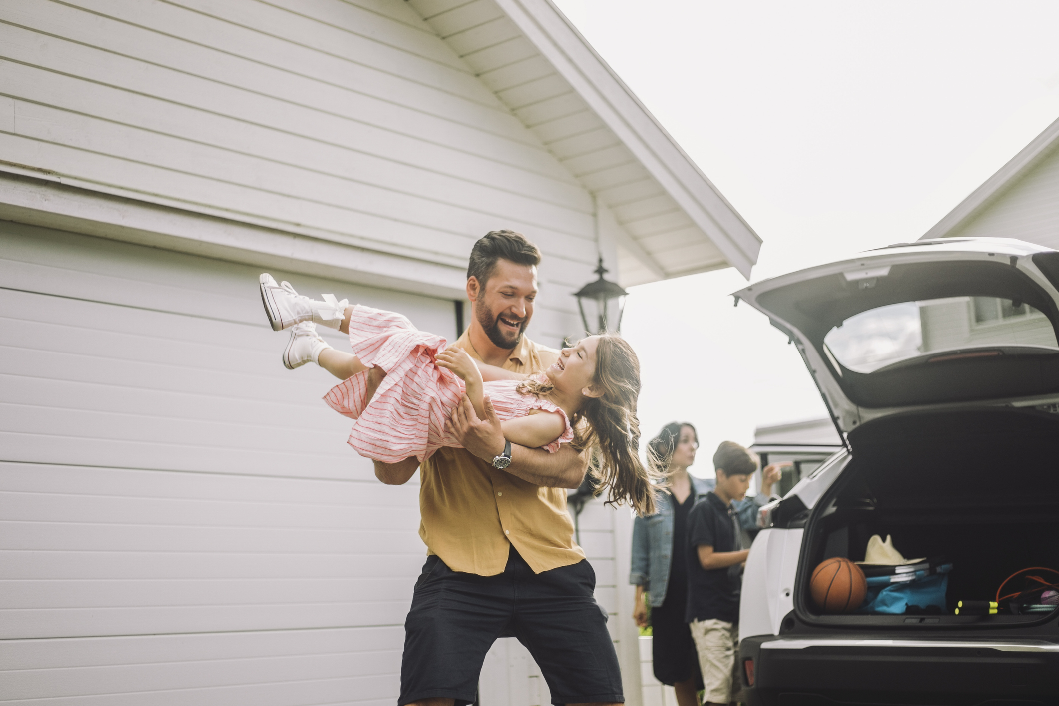 Man lifting a happy child beside an open car trunk at a house, with others nearby