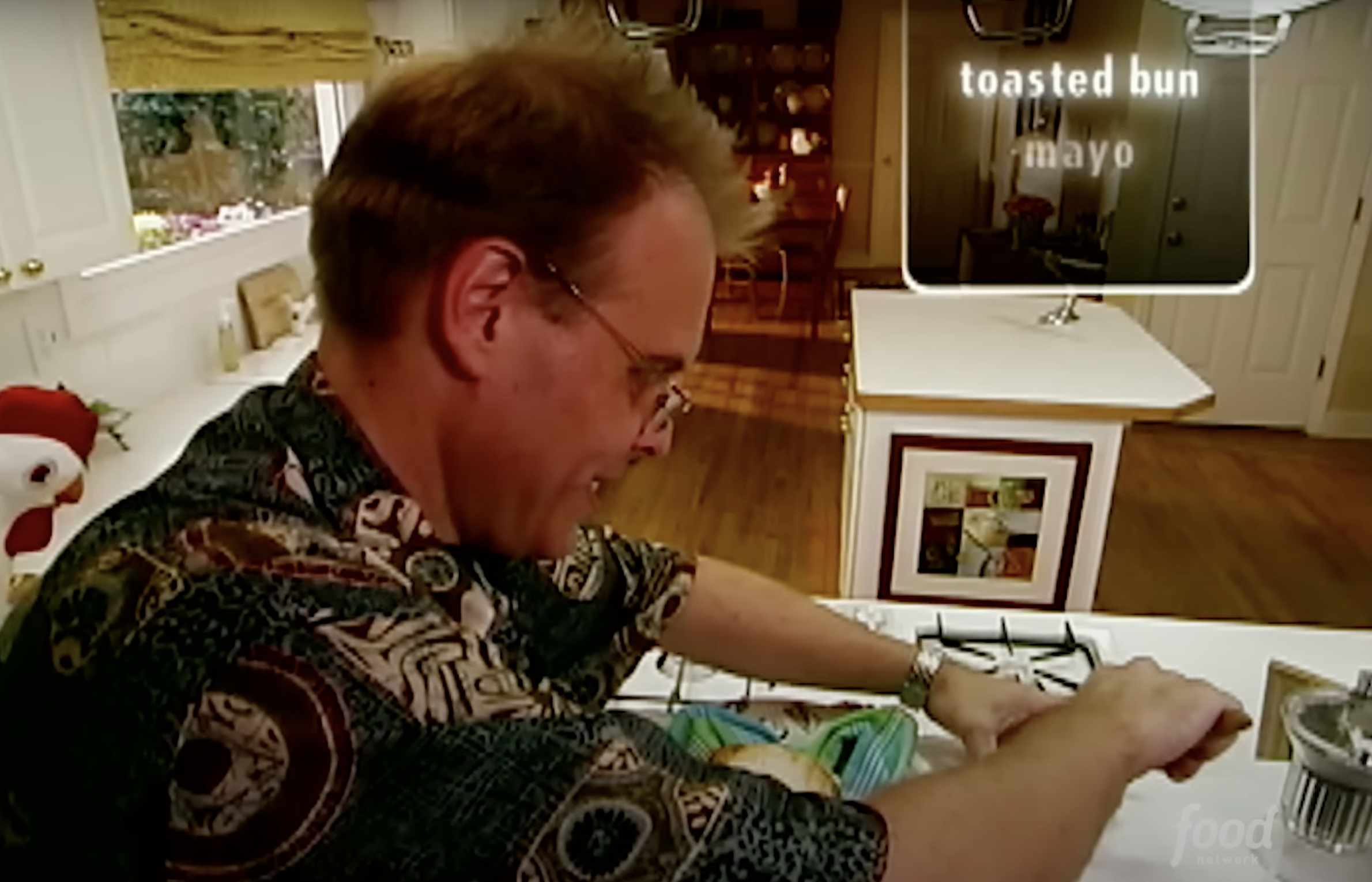 Man in patterned shirt toasting bun, condiments on counter, TV show scene from &quot;Good Eats&quot; with Alton Brown