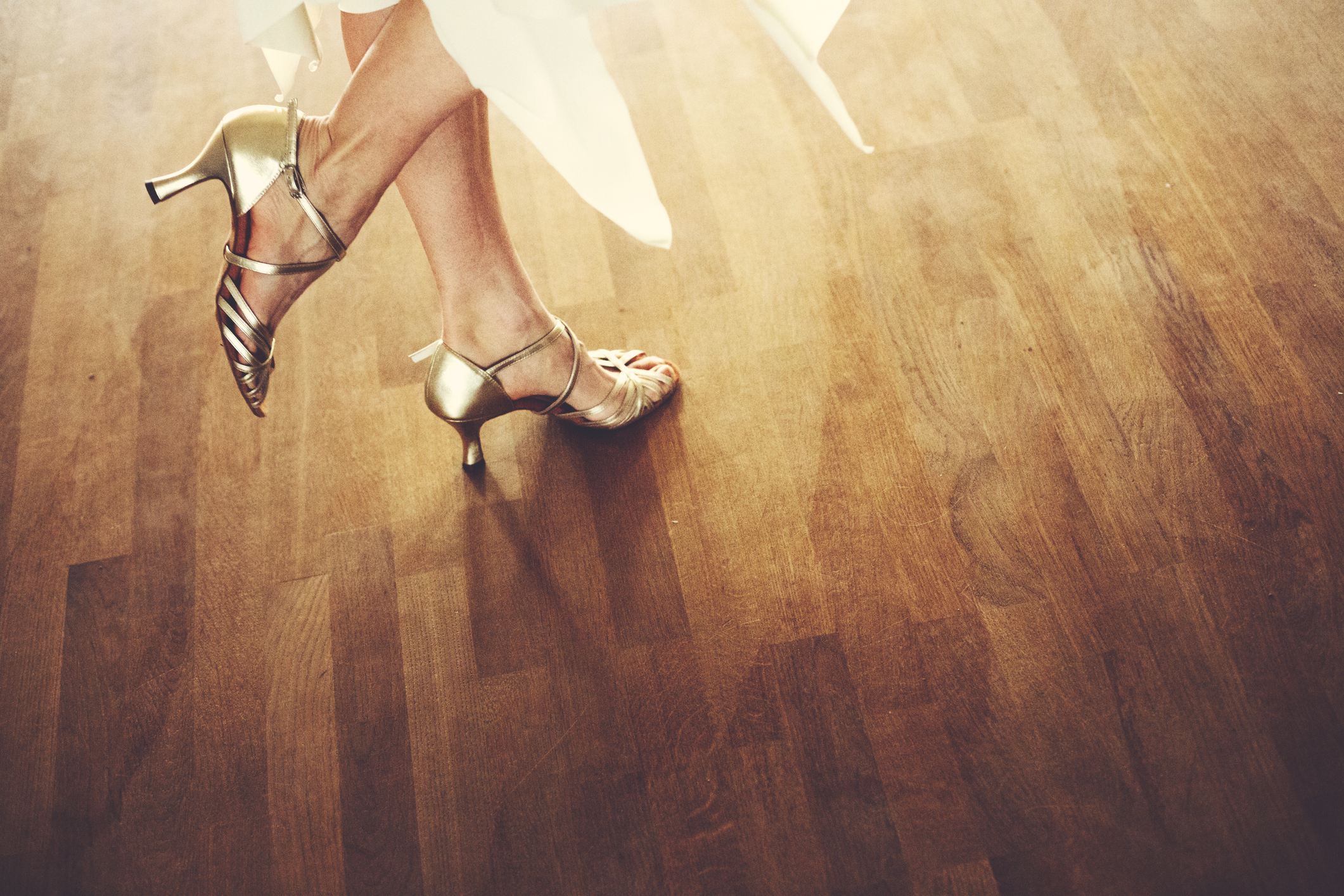 Woman in elegant strappy heels and the hem of a flowing dress, standing poised on a wooden floor
