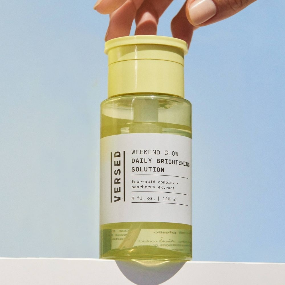 Hand holding Versed Weekend Glow Daily Brightening Solution bottle against a sky backdrop