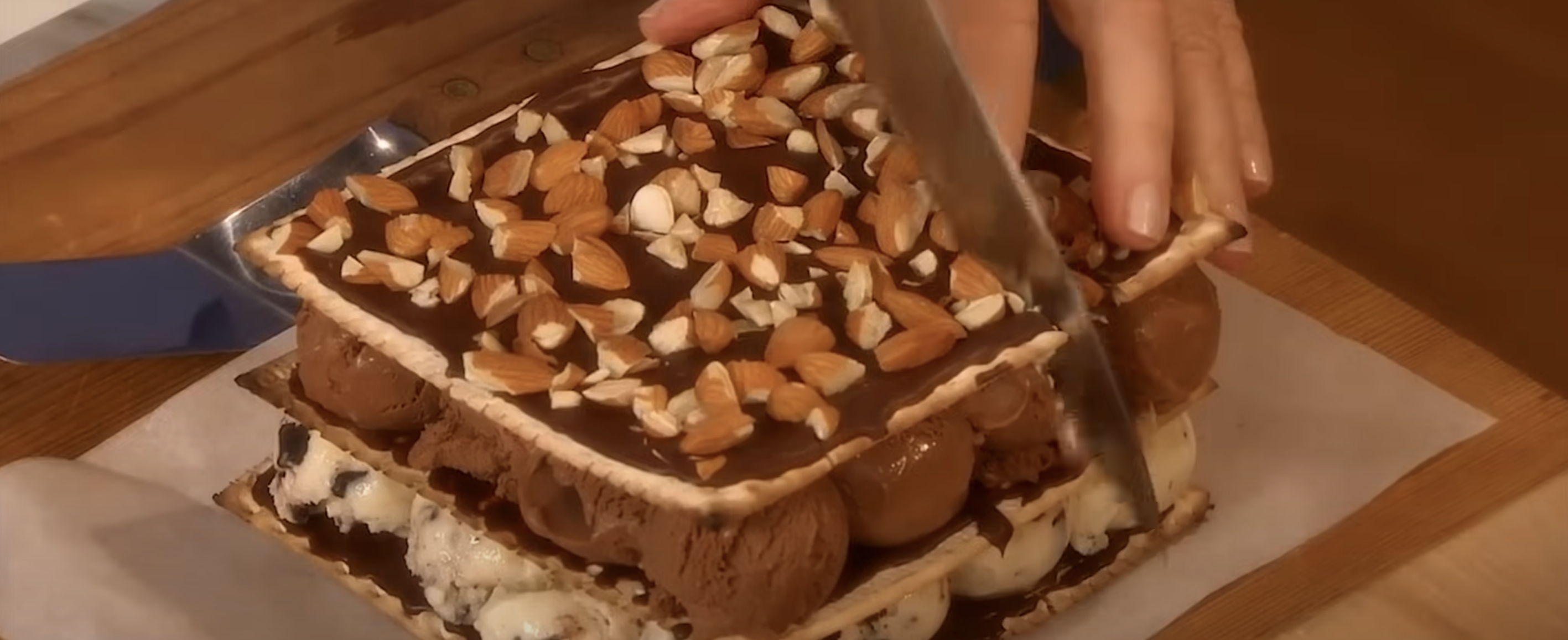 A hand slices a large ice cream sandwich with nuts on top, placed on a wooden board