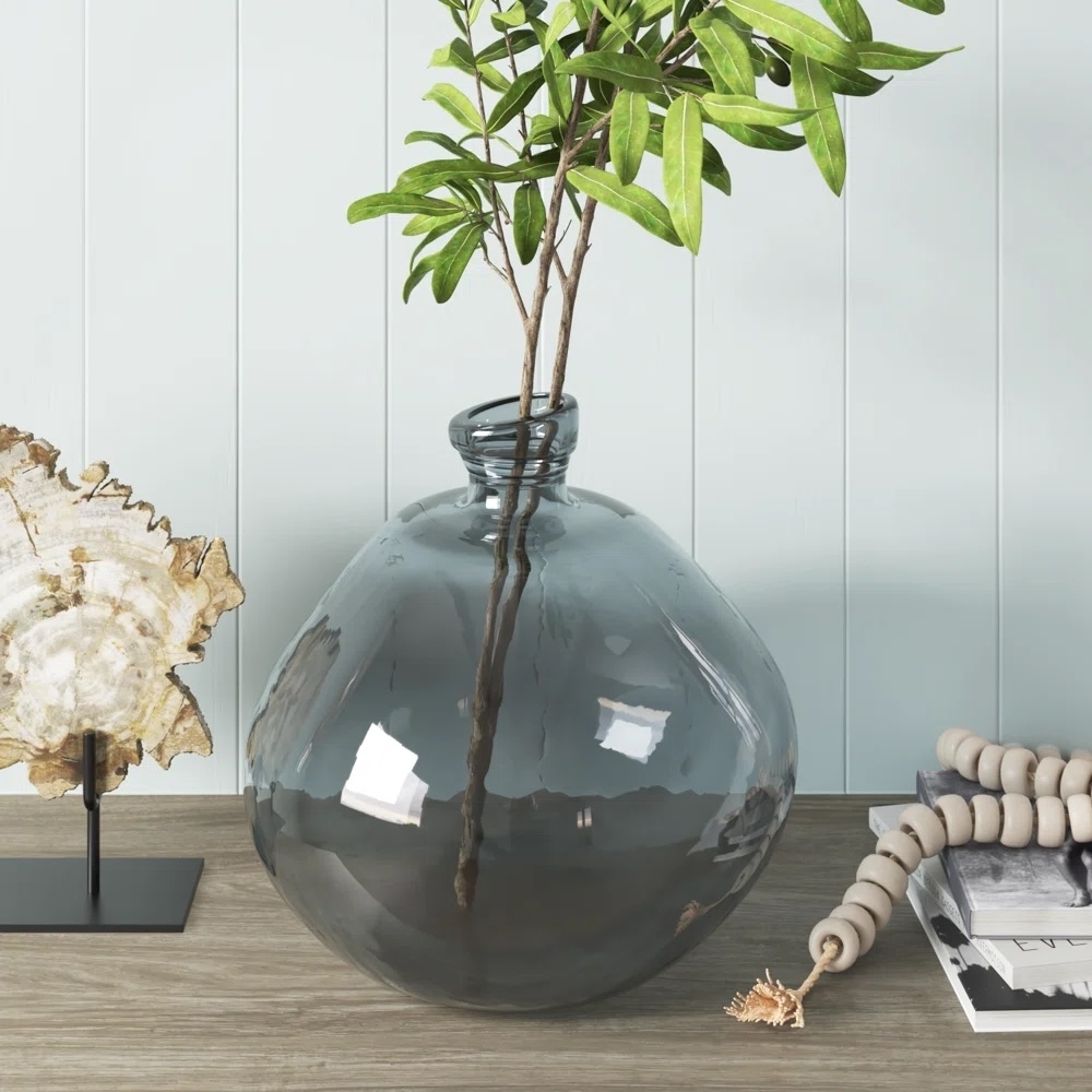 Round glass vase with a plant on a table, beside a book stack and decorative items. Perfect for modern home decor