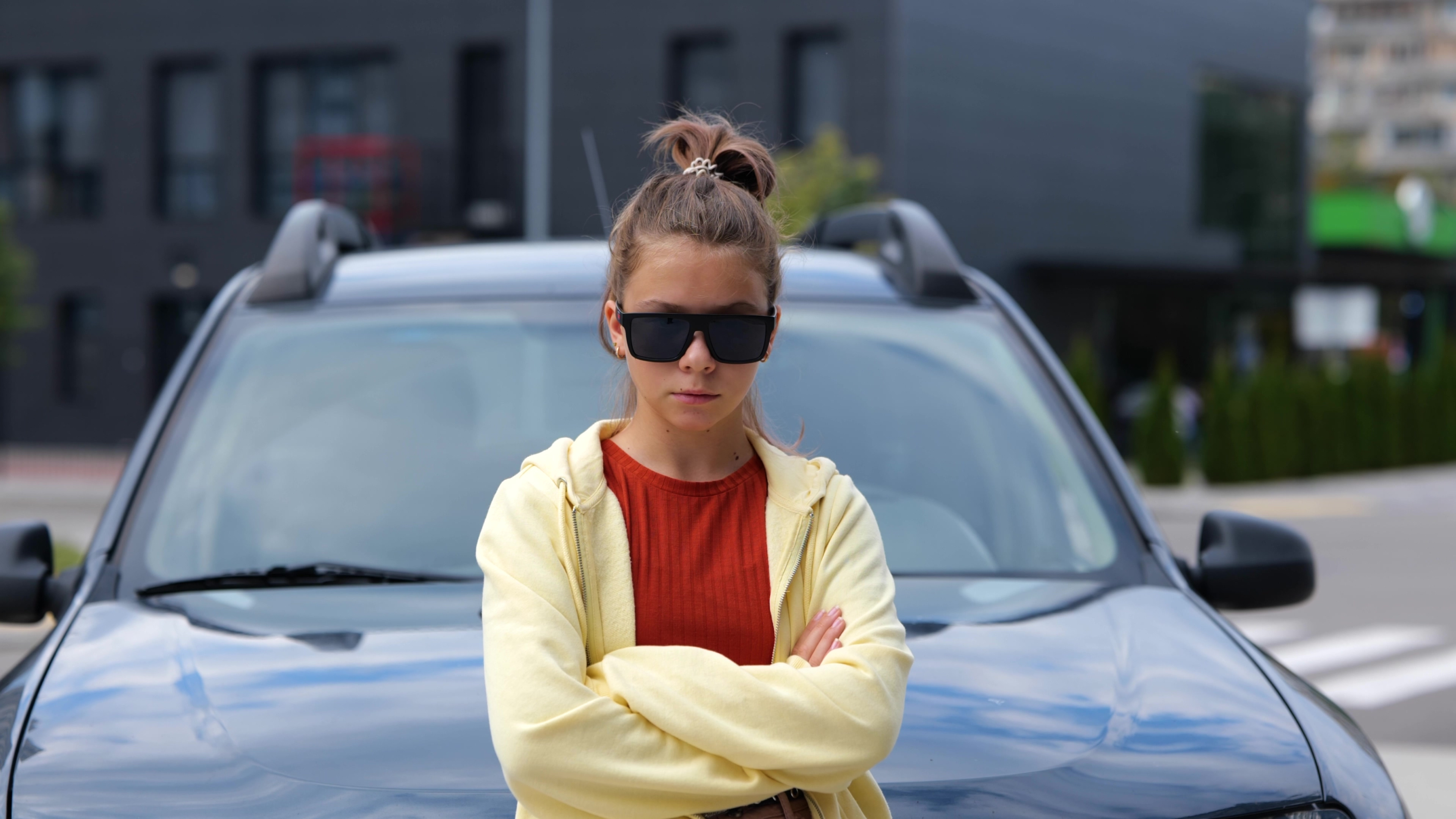 Person with arms crossed standing in front of a car, wearing sunglasses, on a street