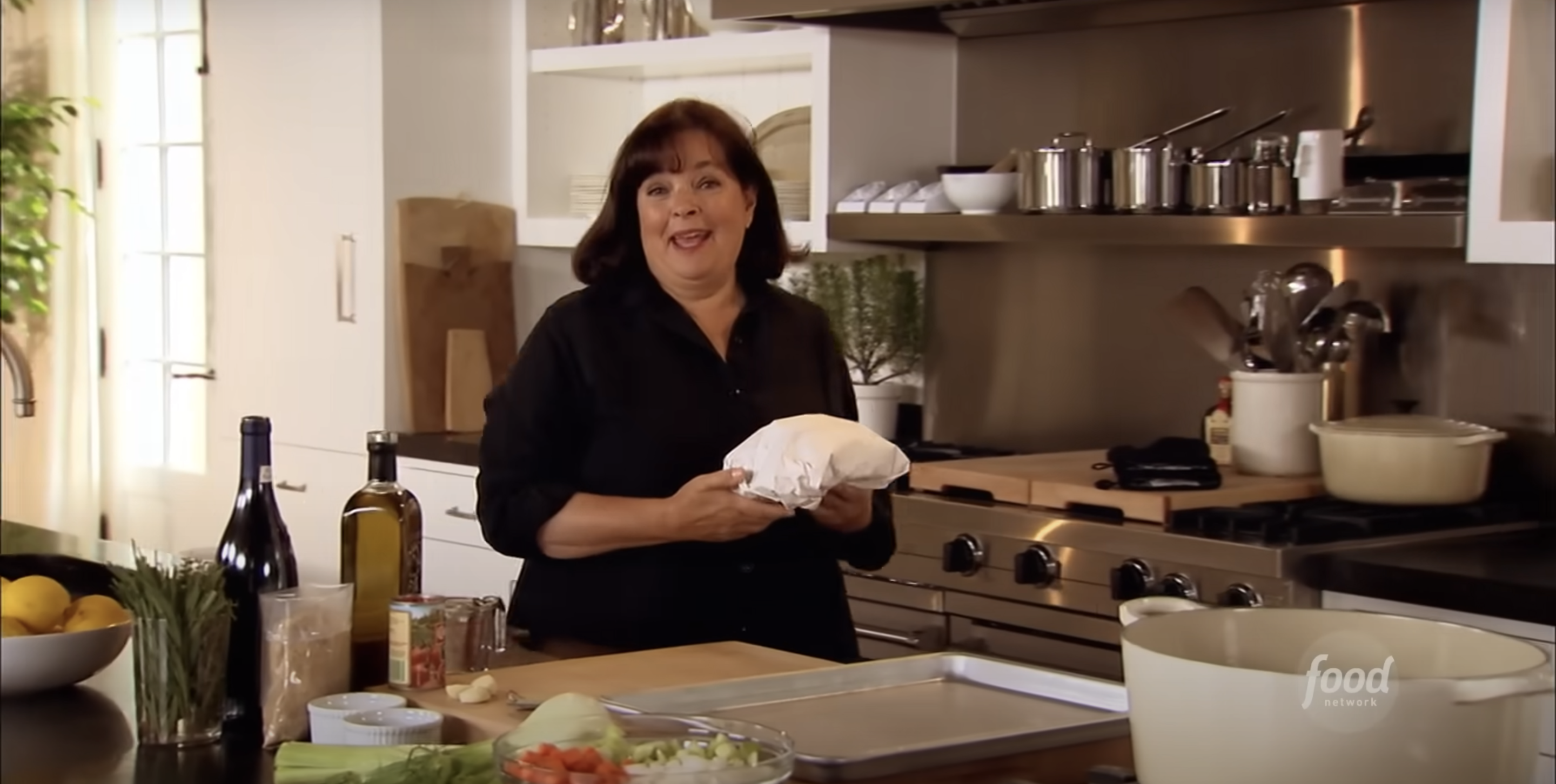 Ina Garten in a kitchen holding a cloth-wrapped item, with ingredients and cookware on the countertop