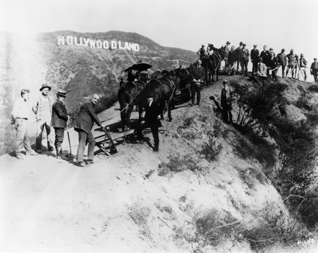 A historical photo of people gathered around a camera and equipment on a hillside with the &quot;Hollywoodland&quot; sign in the background