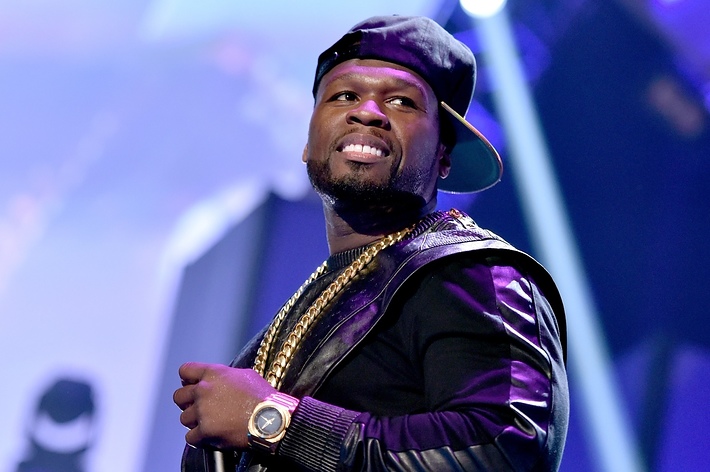 50 Cent wearing a black cap, heavy gold chain, and a purple jacket while performing onstage