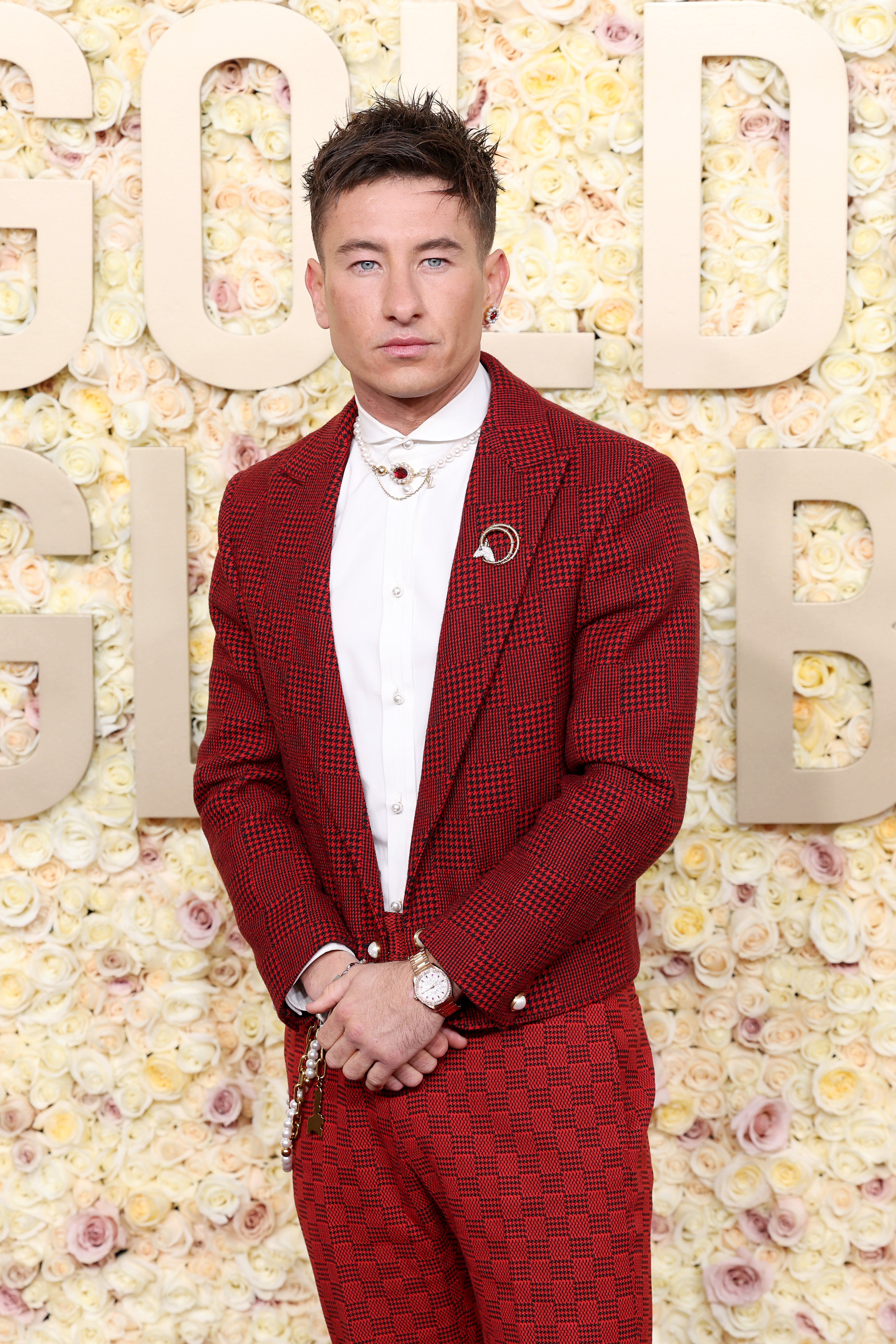Man in a patterned red suit with hands clasped in front of a flower wall with gold letters