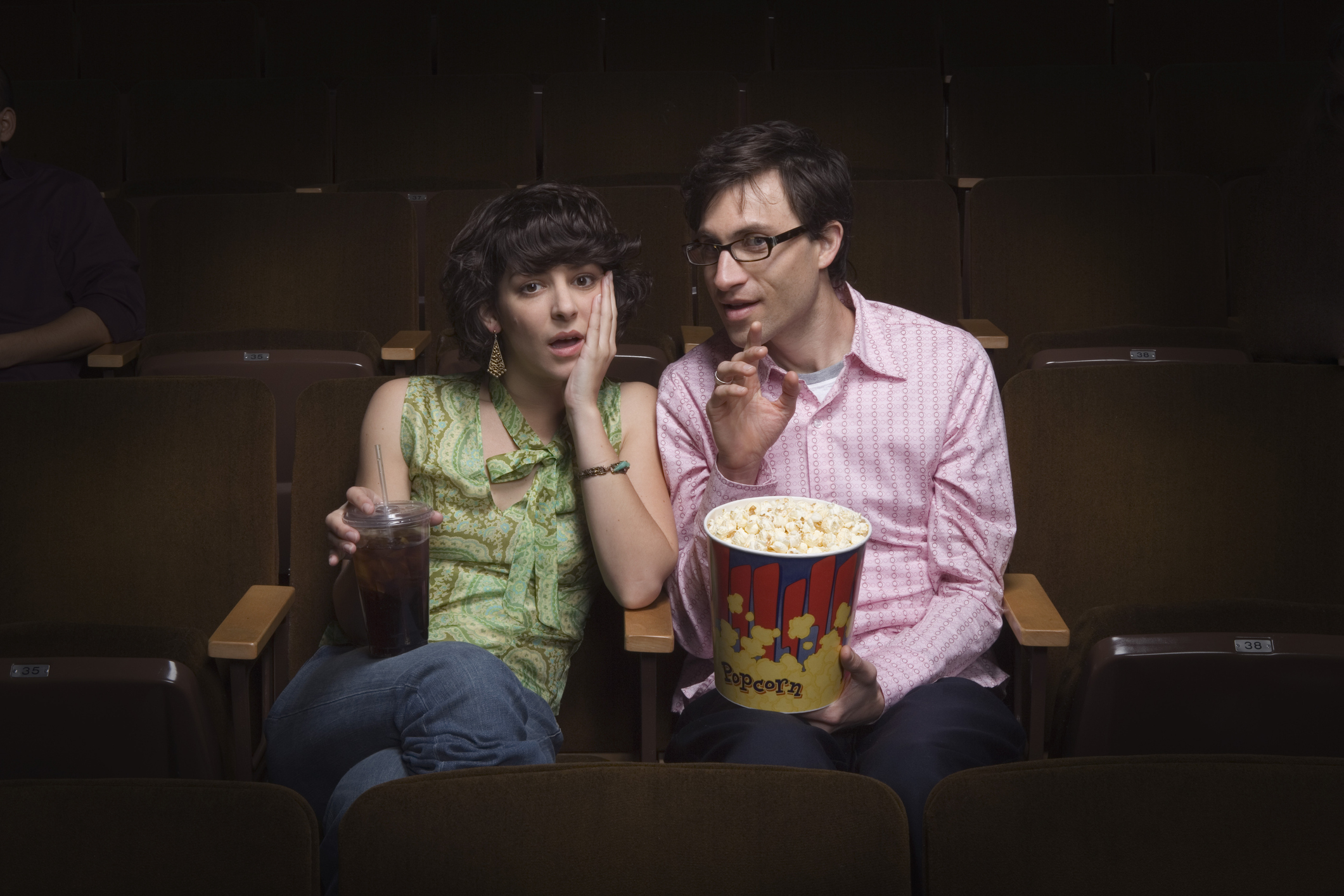 Two people sitting in a theater seem surprised with one whispering to the other; both hold snacks