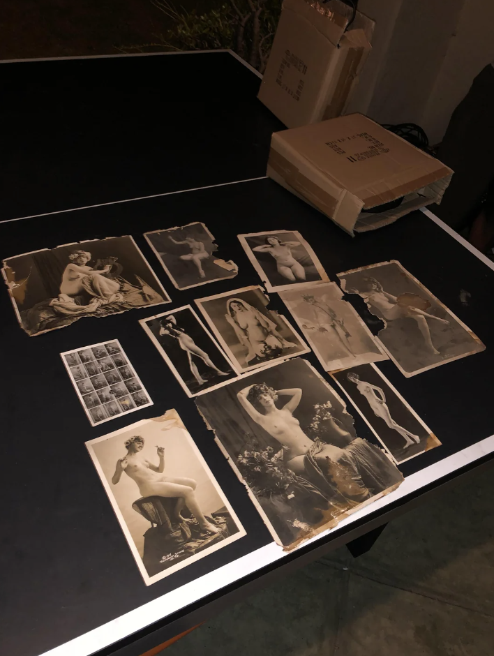 Vintage photos spread on a table, some featuring risque poses, alongside an open book