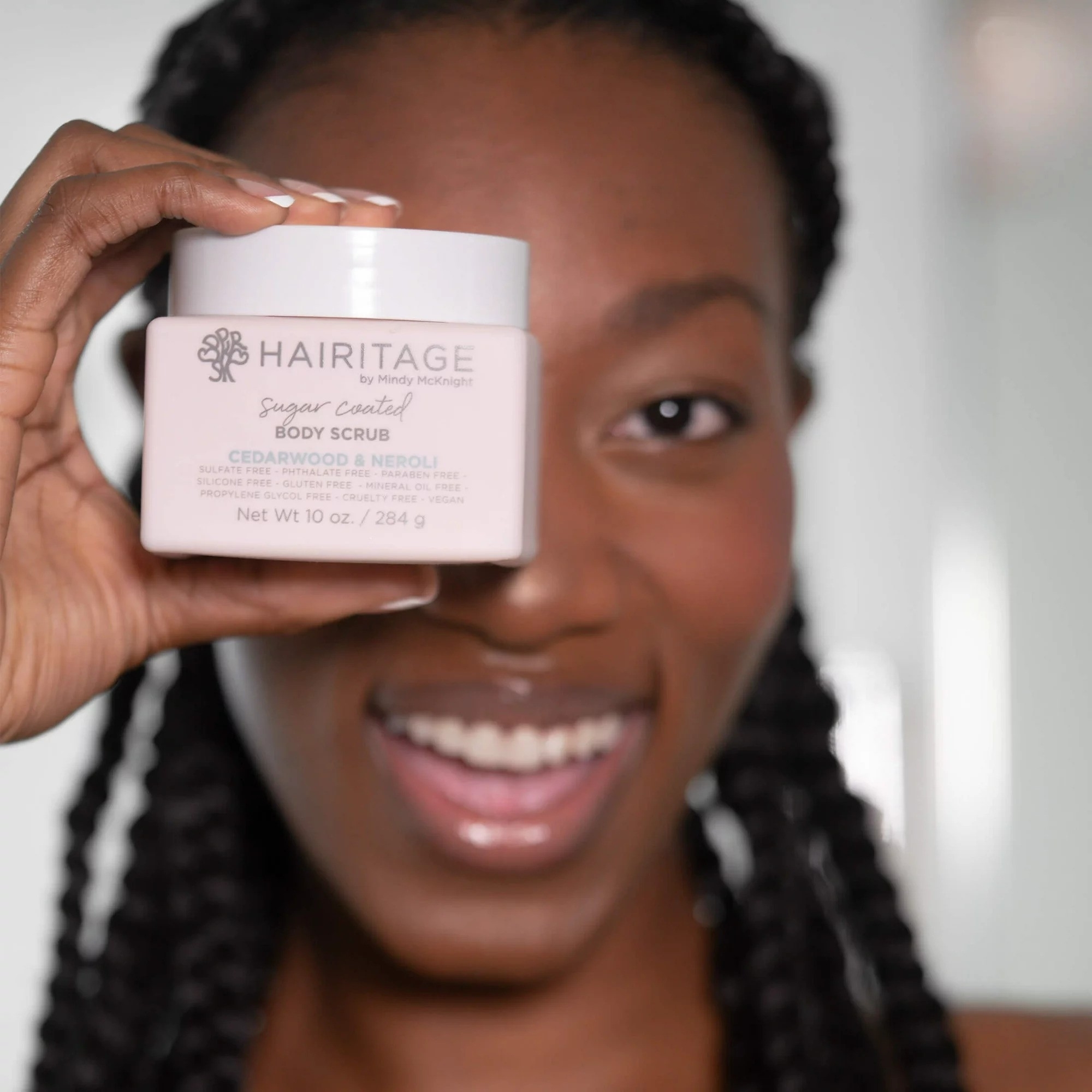 Model smiling, holding a Hairitage Sugar Coated Body Scrub jar close to the camera