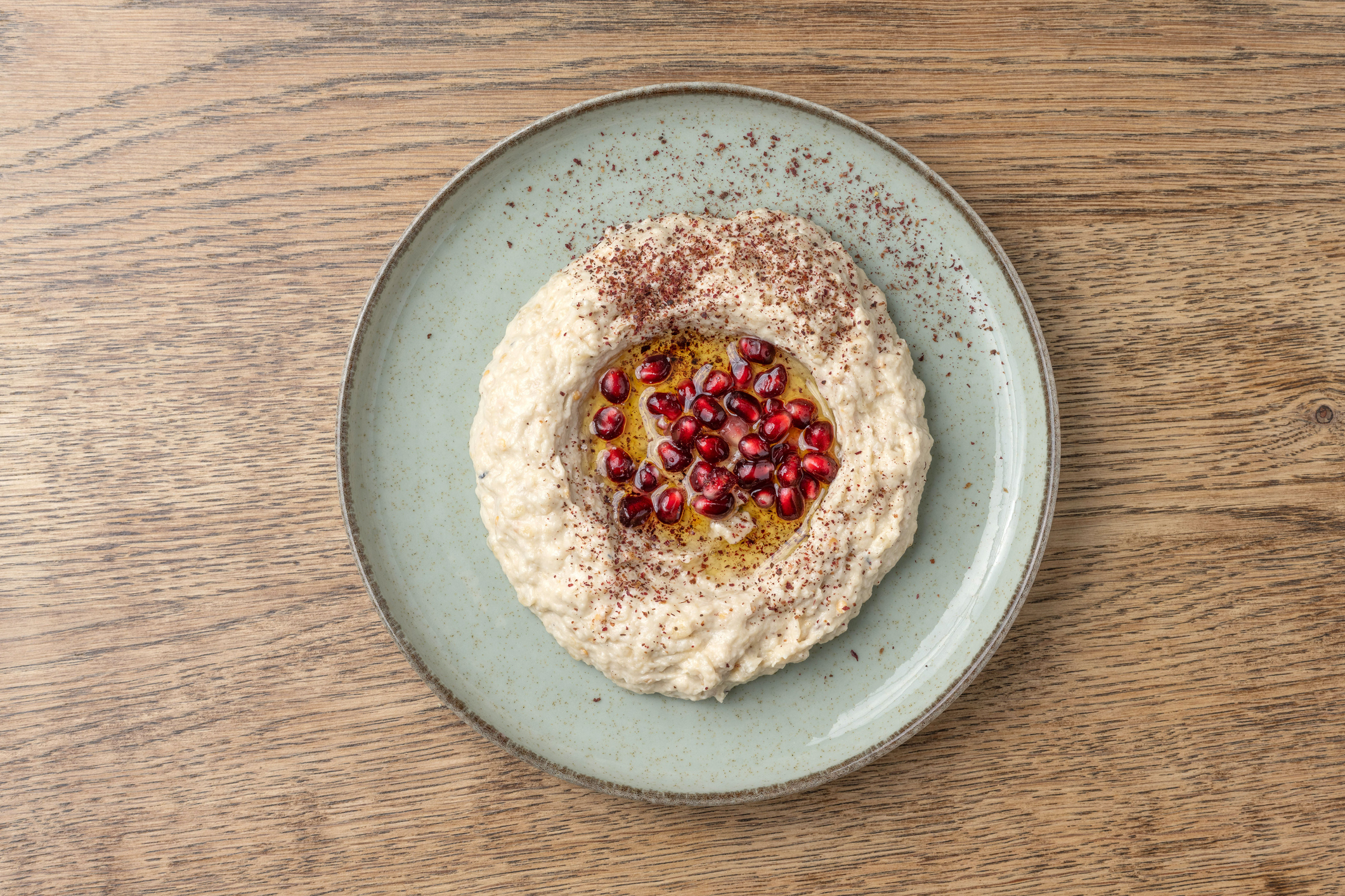 Bowl of baba ghanoush topped with pomegranate seeds and olive oil on a plate, viewed from above