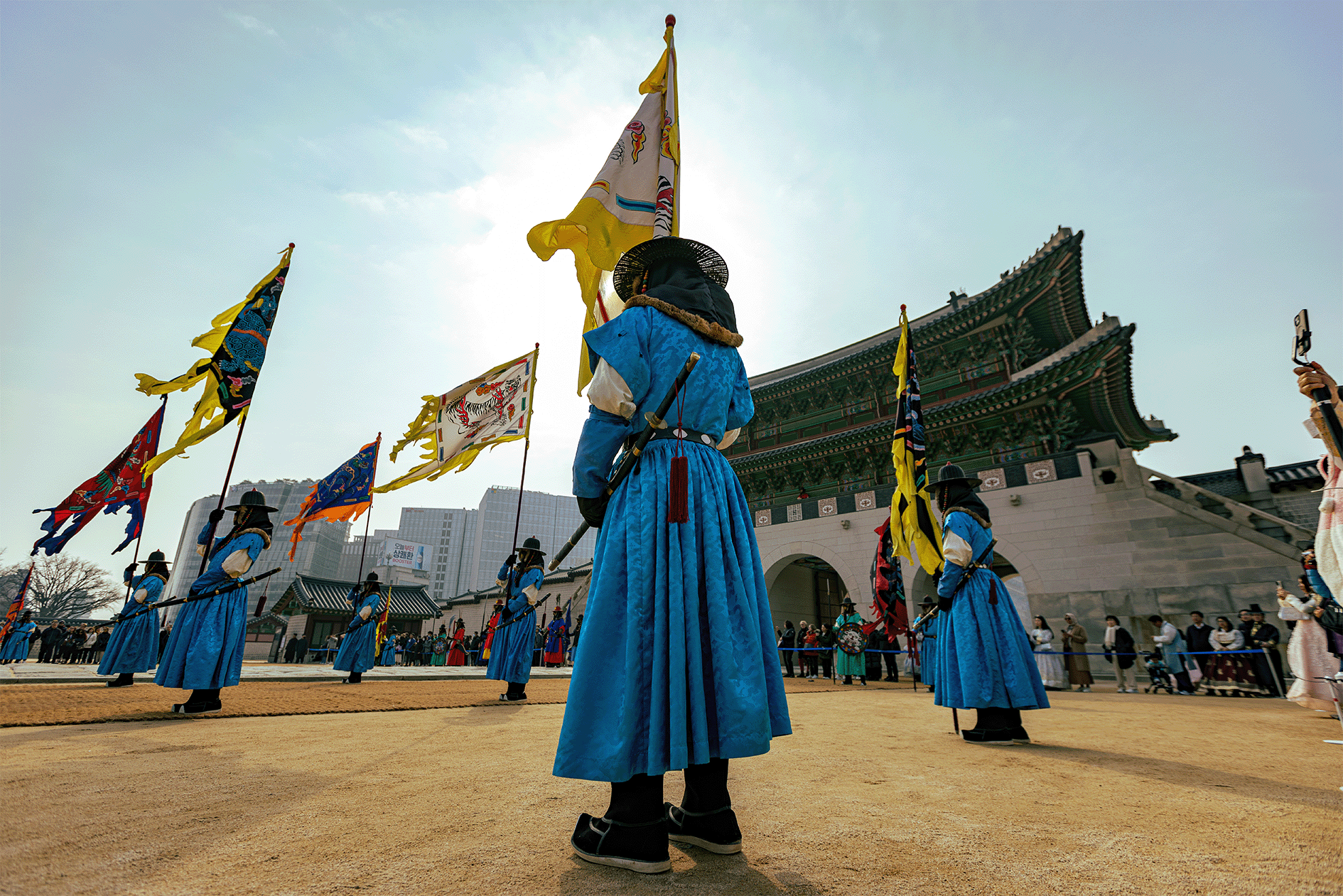 Traditional Korean ceremony with performers in historical attire carrying flags in front of a palace
