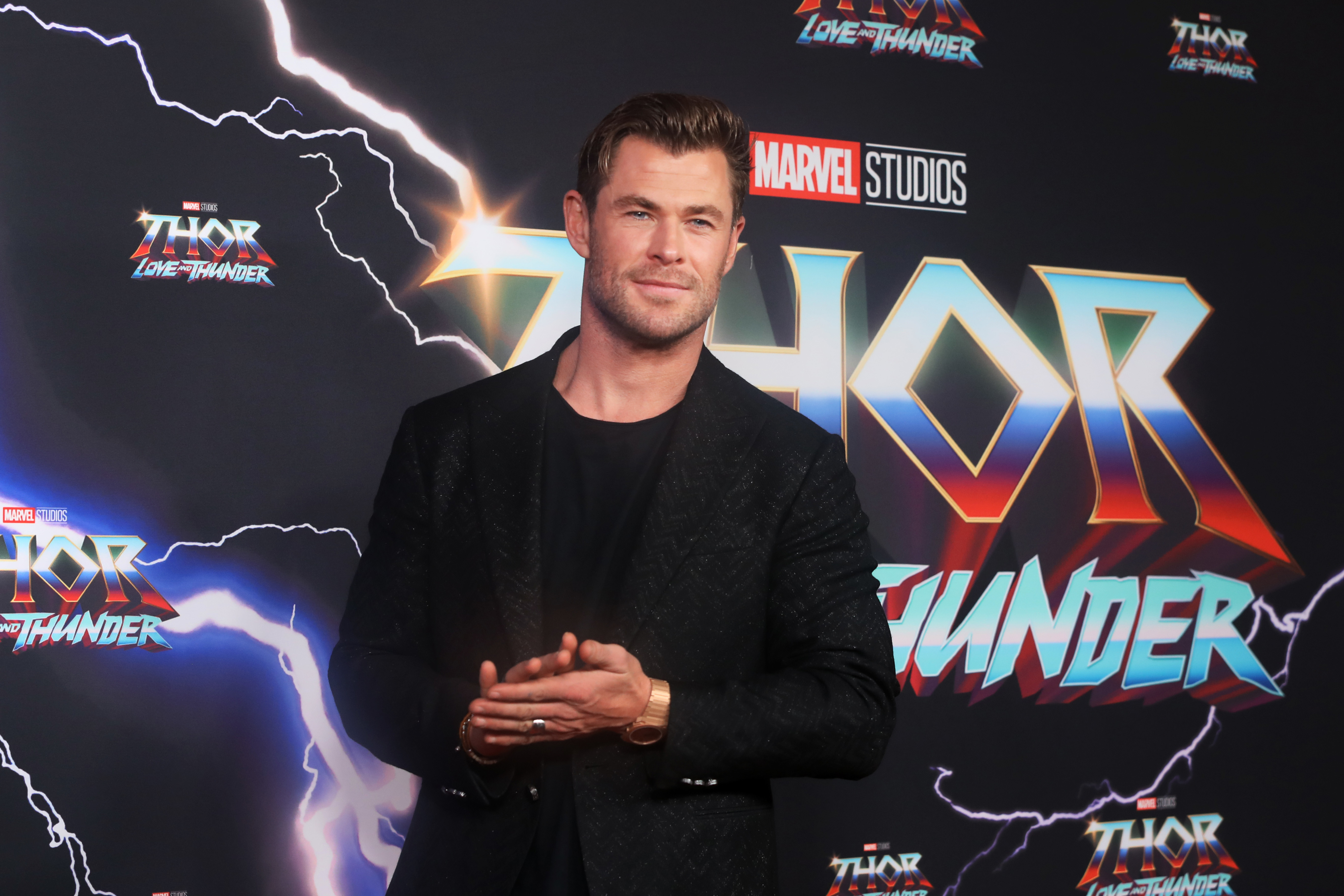 Chris Hemsworth poses at a Thor: Love and Thunder event in a black patterned suit with arms crossed