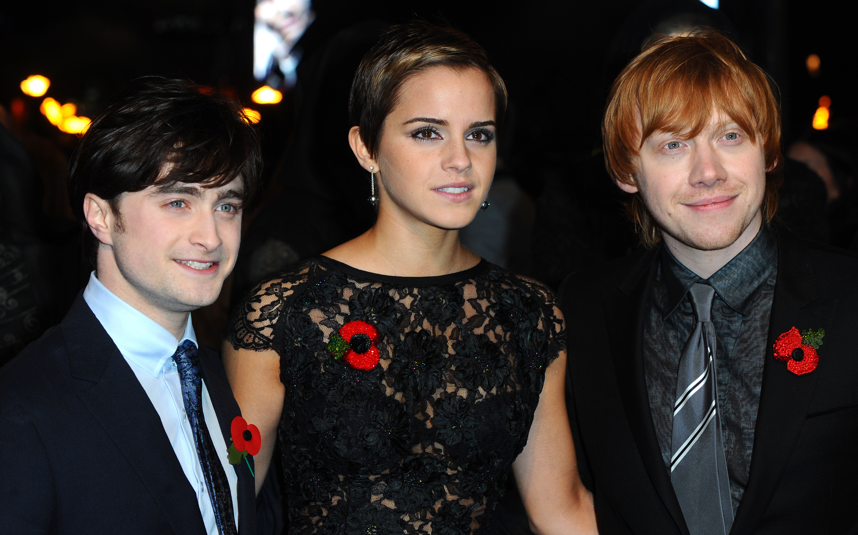 Daniel Radcliffe, Emma Watson, and Rupert Grint smiling at an event, wearing poppy pins. Emma in a lace dress, the men in suits