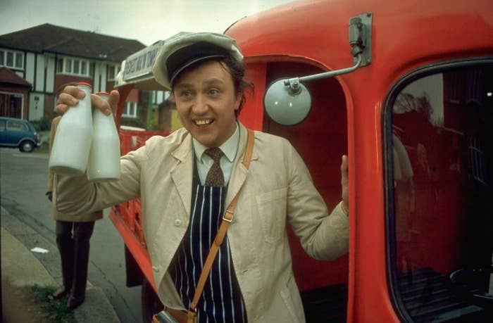 Man in a cap and striped apron holding milk bottles next to a red milk float